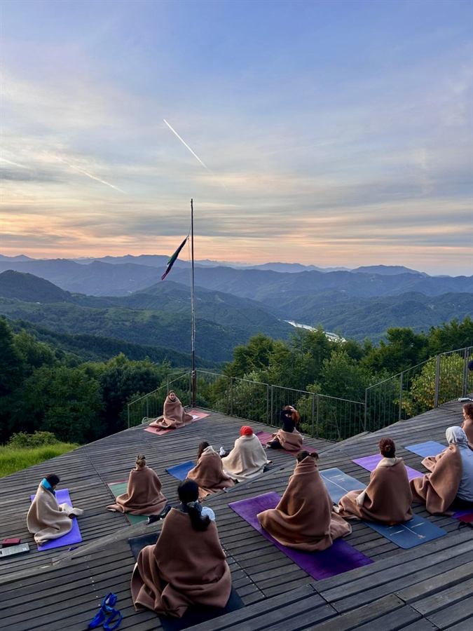 Un gruppo di persone avvolte in coperte, sedute sui tappetini su una terrazza di legno affacciata su valli e montagne, sotto un cielo rosato